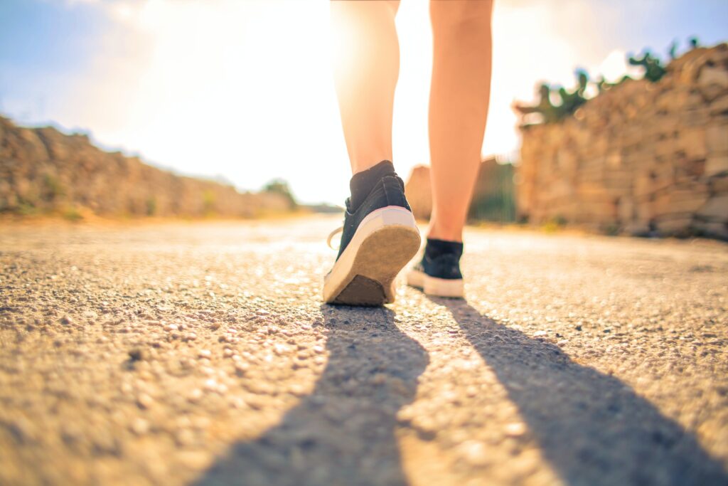 Image d'une personne qui marche sur du bitume vers le soleil, ne voyant que les jambes et pieds. Article sur la marche sur le blog Serenity Body Mind. #SBM #marche